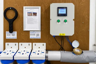 A control panel on a plywood wall beside framed commissioning notices, a water meter and a tank float.