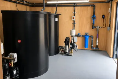 Two black settling tanks and a pump set in a plywood-lined plant room.