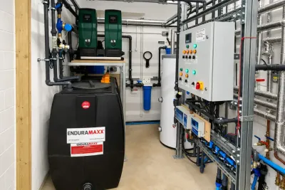 A plant room with a black Enduramaxx storage tank, a wall-mounted control panel, twin pumps and labelled pipework.