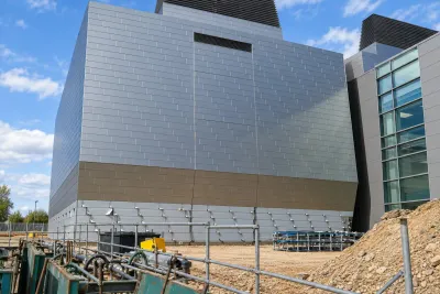A large grey-clad commercial building under construction, with pipework in the trench at its base.