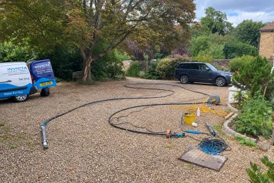 Pipework and hoses laid across a gravel drive, with an Invicta van and a car under the trees.