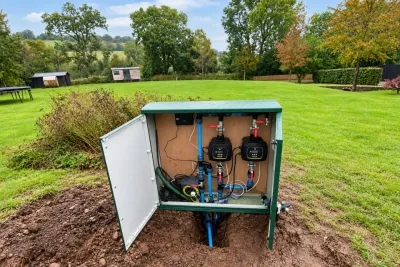 A green borehole kiosk in a field with its doors open, showing the pump controllers and valves inside.