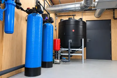 A plant room with three blue pressure vessels, a black storage tank and a grey steel door.